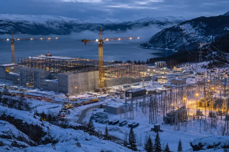 Steel framed AI data center under construction beside a snow covered Norwegian fjord at dusk, with two tower cranes and a lit electrical substation and transmission lines