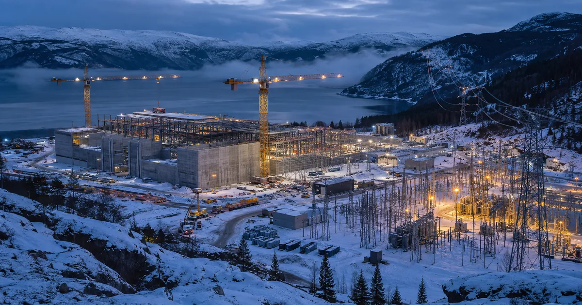 Steel framed AI data center under construction beside a snow covered Norwegian fjord at dusk, with two tower cranes and a lit electrical substation and transmission lines